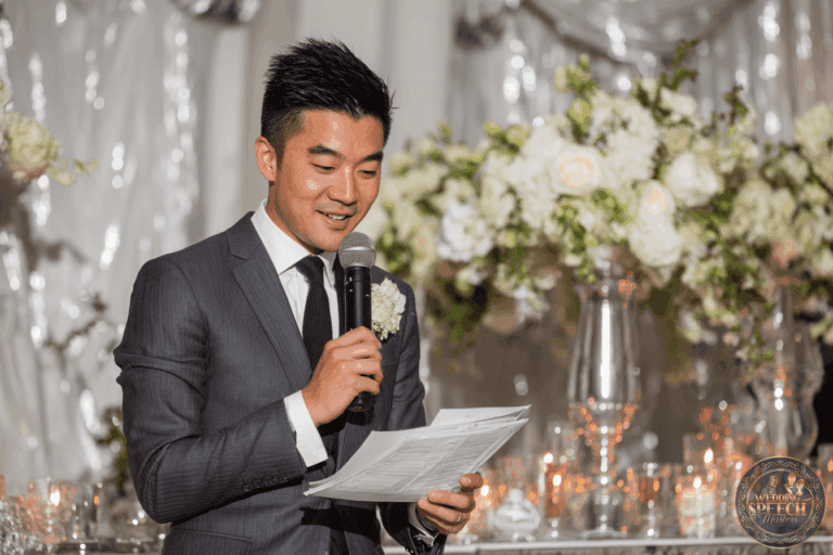 A man in a suit speaks into a microphone while holding papers, standing in front of a decorated table with white flowers and candles.