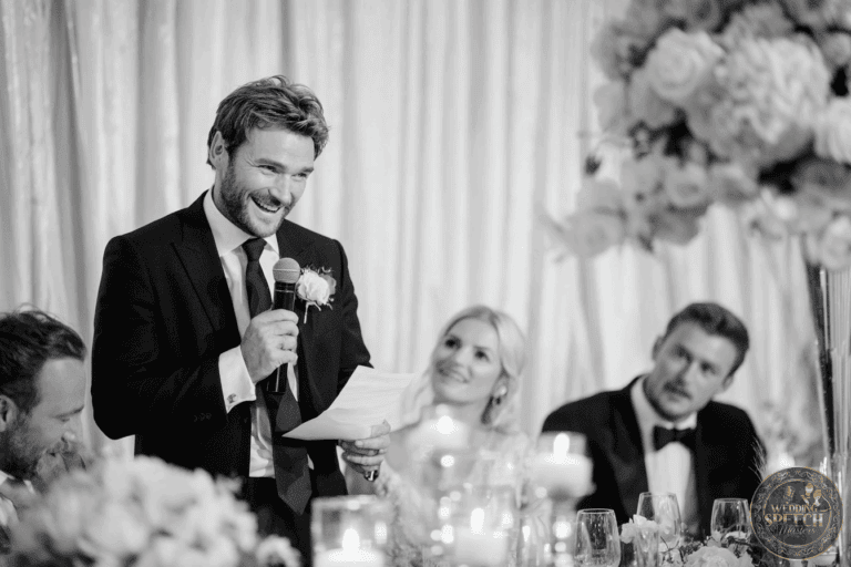 A man in a suit speaks into a microphone while holding a paper at a formal event, with seated guests and floral decorations nearby.