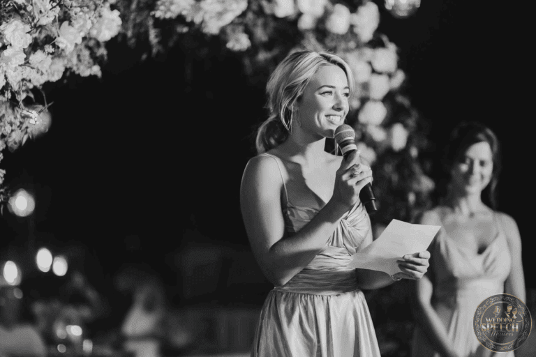A woman in a formal dress holds a microphone and a piece of paper, speaking at an event. Another woman stands in the background.