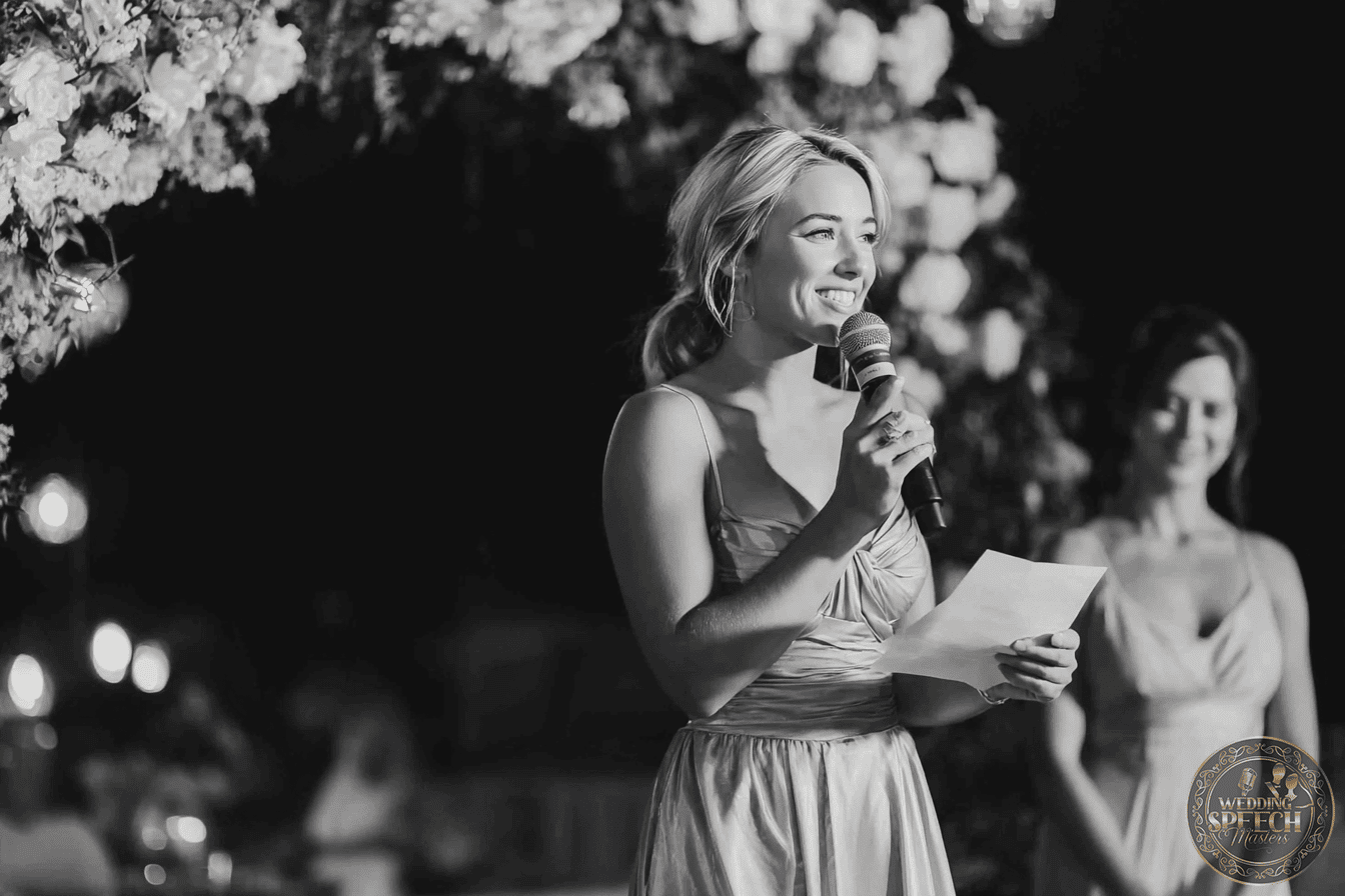 A woman in a formal dress holds a microphone and a piece of paper, speaking at an event. Another woman stands in the background.