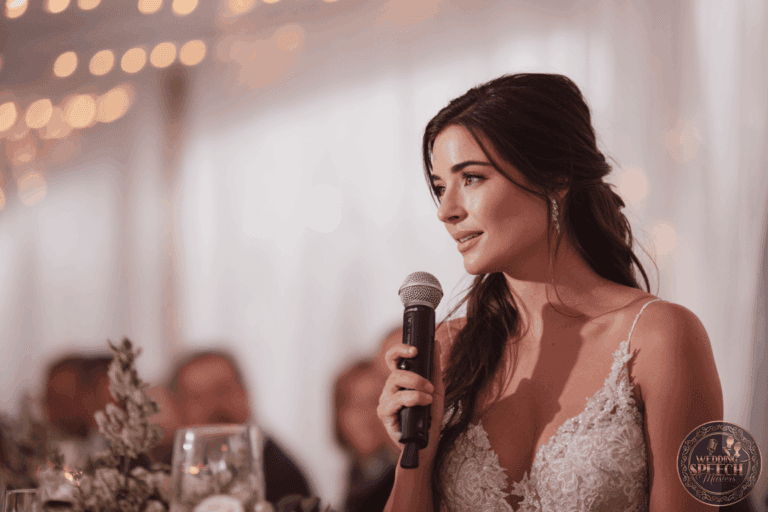 A woman in a light lace dress holds a microphone and speaks at an indoor event, with blurred people and string lights in the background.