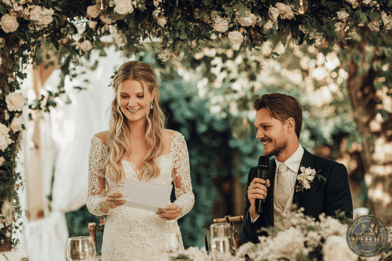 A bride in a lace wedding dress reads from a paper while a groom in a suit with a boutonniere holds a microphone, both seated under a floral arch at an outdoor wedding.