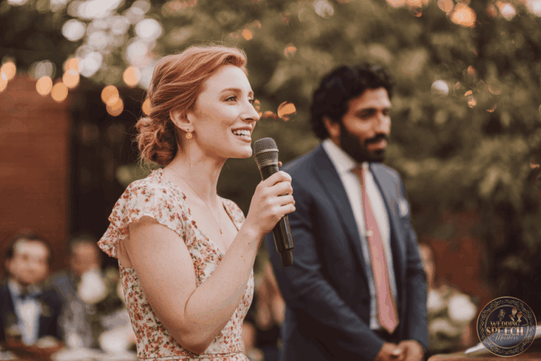 A woman in a floral dress speaks into a microphone at an outdoor event, with a man in a suit standing beside her and blurred guests in the background.
