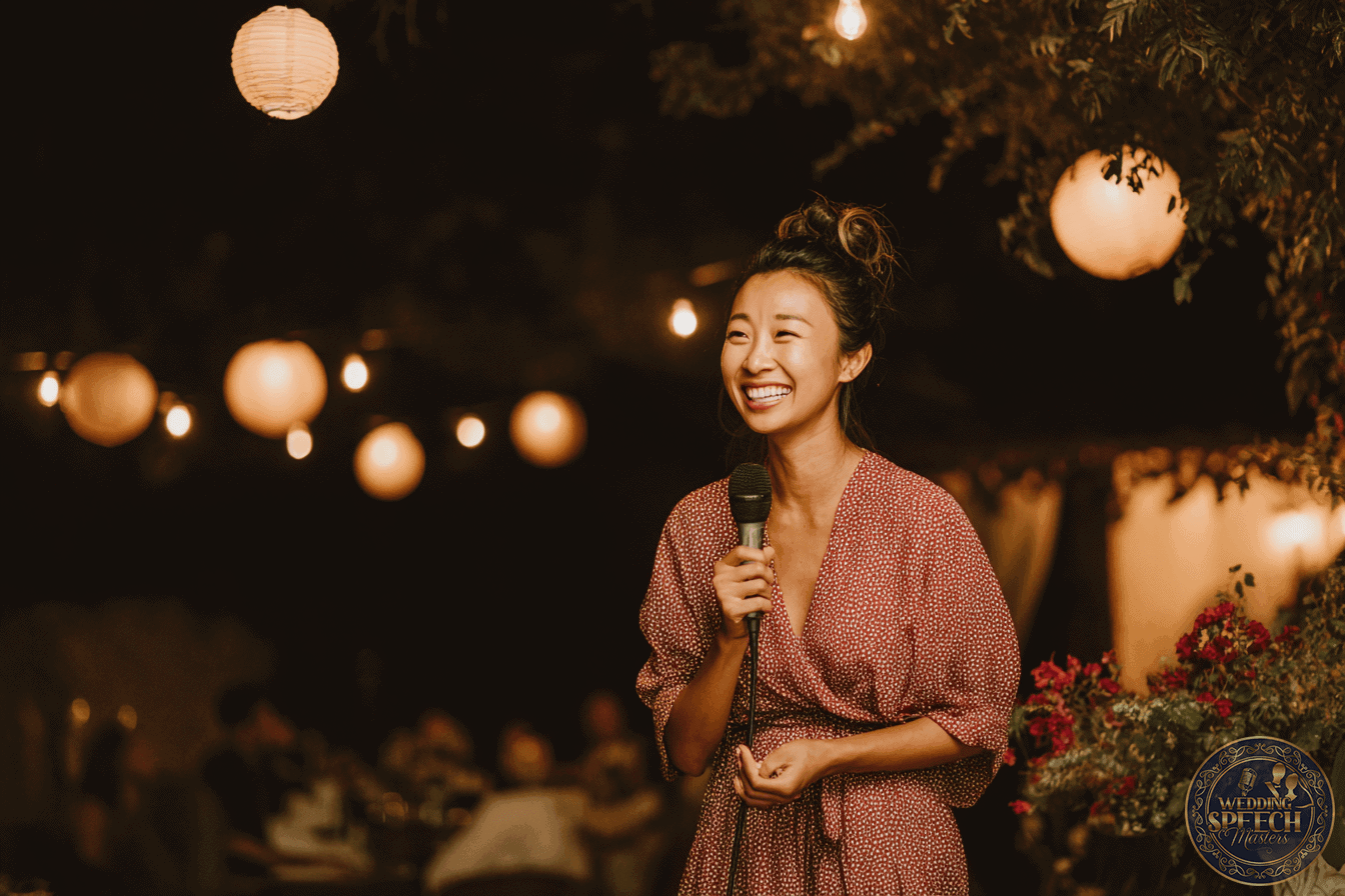 A woman in a patterned dress smiles while holding a microphone at an outdoor event with string lights and lanterns in the background.