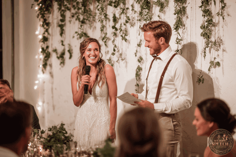 A bride in a white dress smiles while speaking into a microphone next to a man holding papers at a decorated wedding reception.