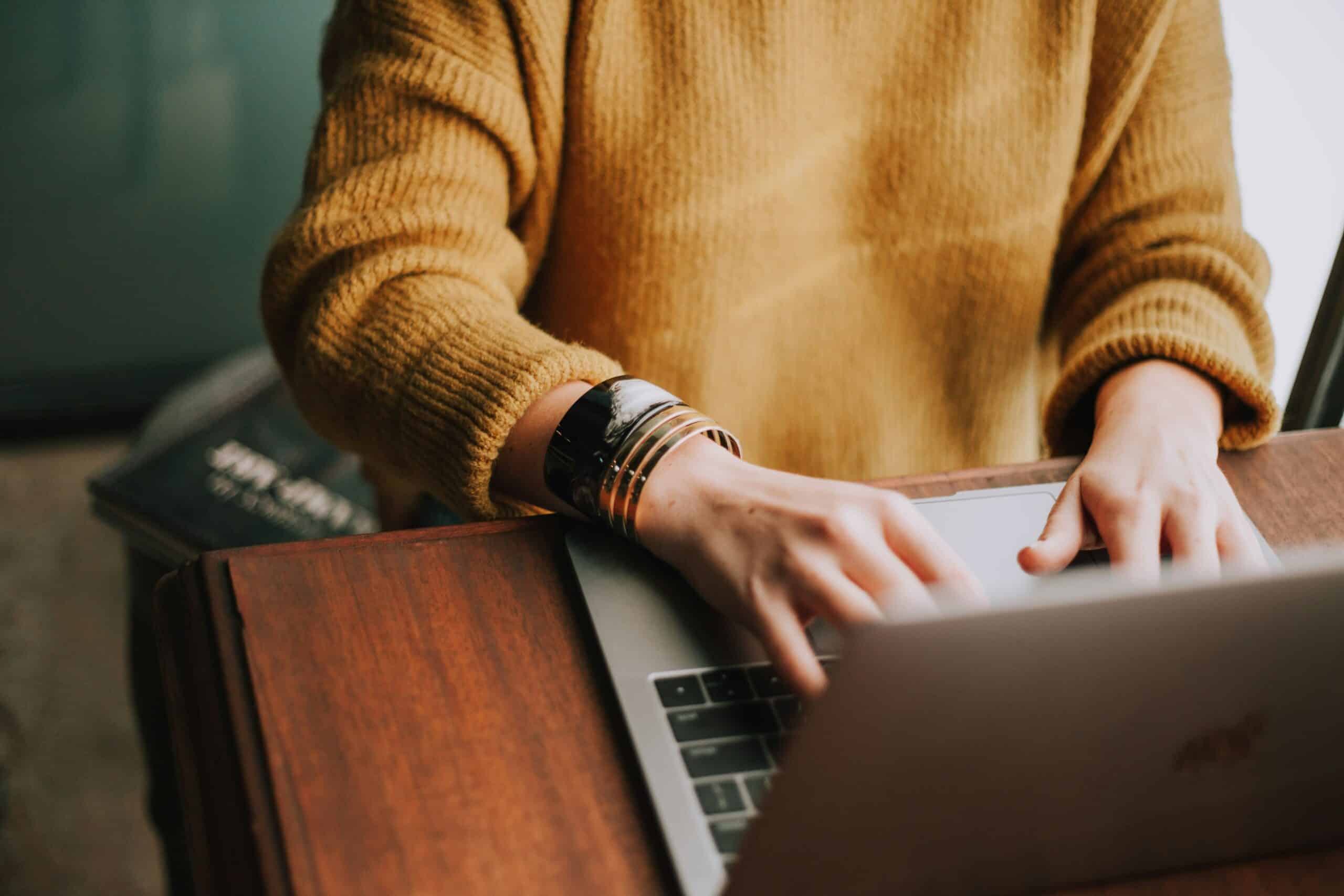 Person in a yellow sweater sitting at a table typing on a laptop.
