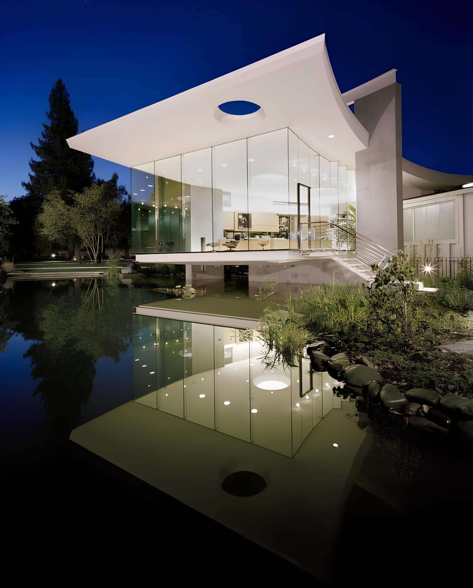 Modern contemporary house with sleek white architecture and glass exterior reflecting water feature, situated in lush green landscape at night.