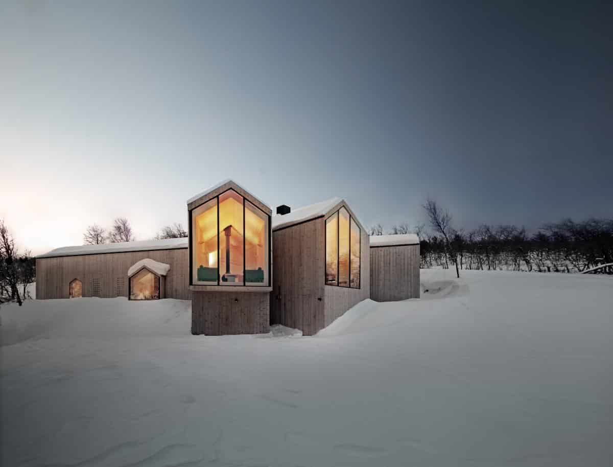 Modern snow-covered house with contemporary architecture, large glass windows, and wooden exterior in a winter landscape.