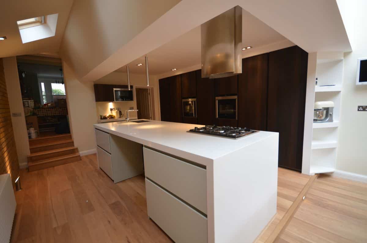 Sleek modern kitchen with white island and dark wood cabinets, featuring built-in appliances and stainless steel range hood, illuminated by recessed lighting and natural skylights for contemporary architectural design.
