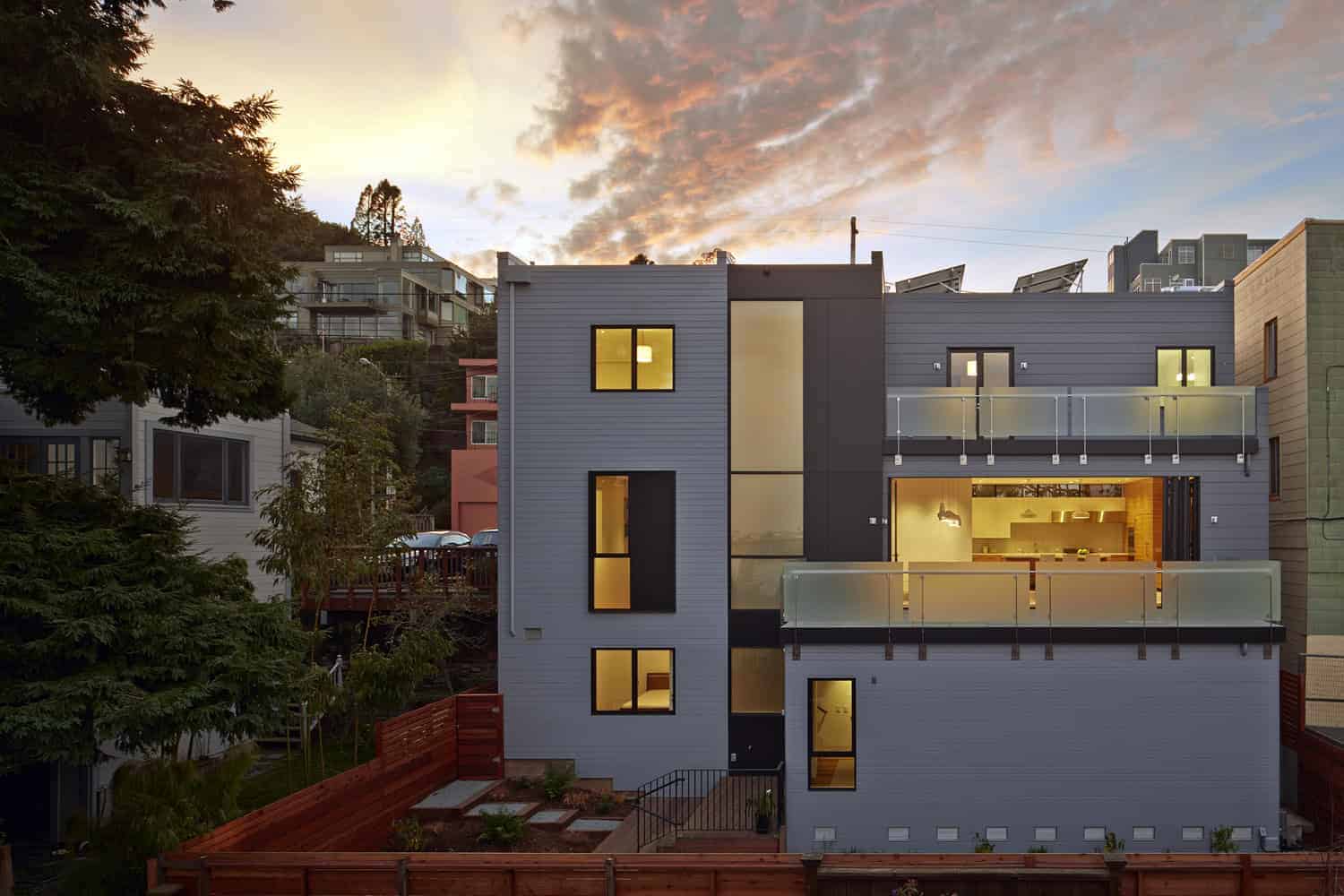 Sleek modern multi-story house with contemporary architecture, large windows, and spacious balconies set against a scenic hillside backdrop during sunset.