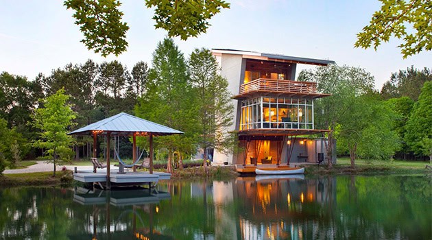 Modern lakeside house with large glass windows and surrounding lush greenery, showcasing contemporary architecture and scenic water views.