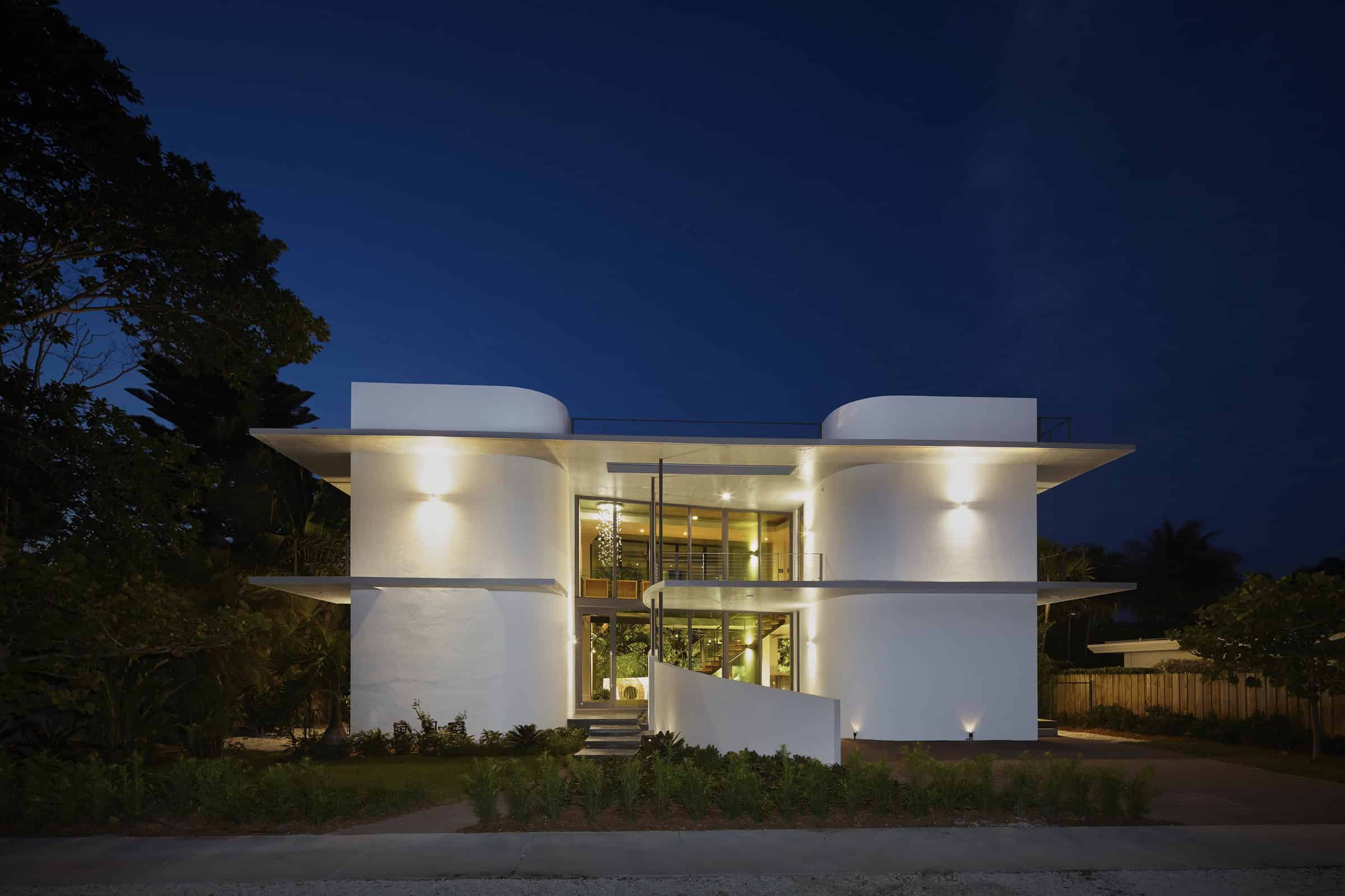 Modern white architectural house with curved walls and large glass windows at night, featuring sleek lighting and contemporary design elements.
