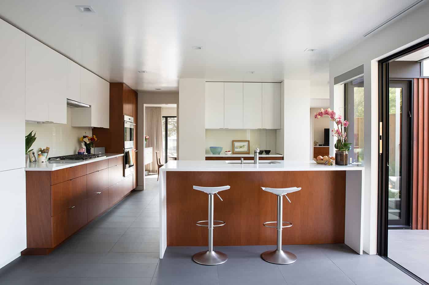 Sleek modern kitchen with minimalist design, wooden cabinetry, white countertops, and open layout, featuring a kitchen island, modern bar stools, and large sliding glass doors, perfect for contemporary architecture art designs.
