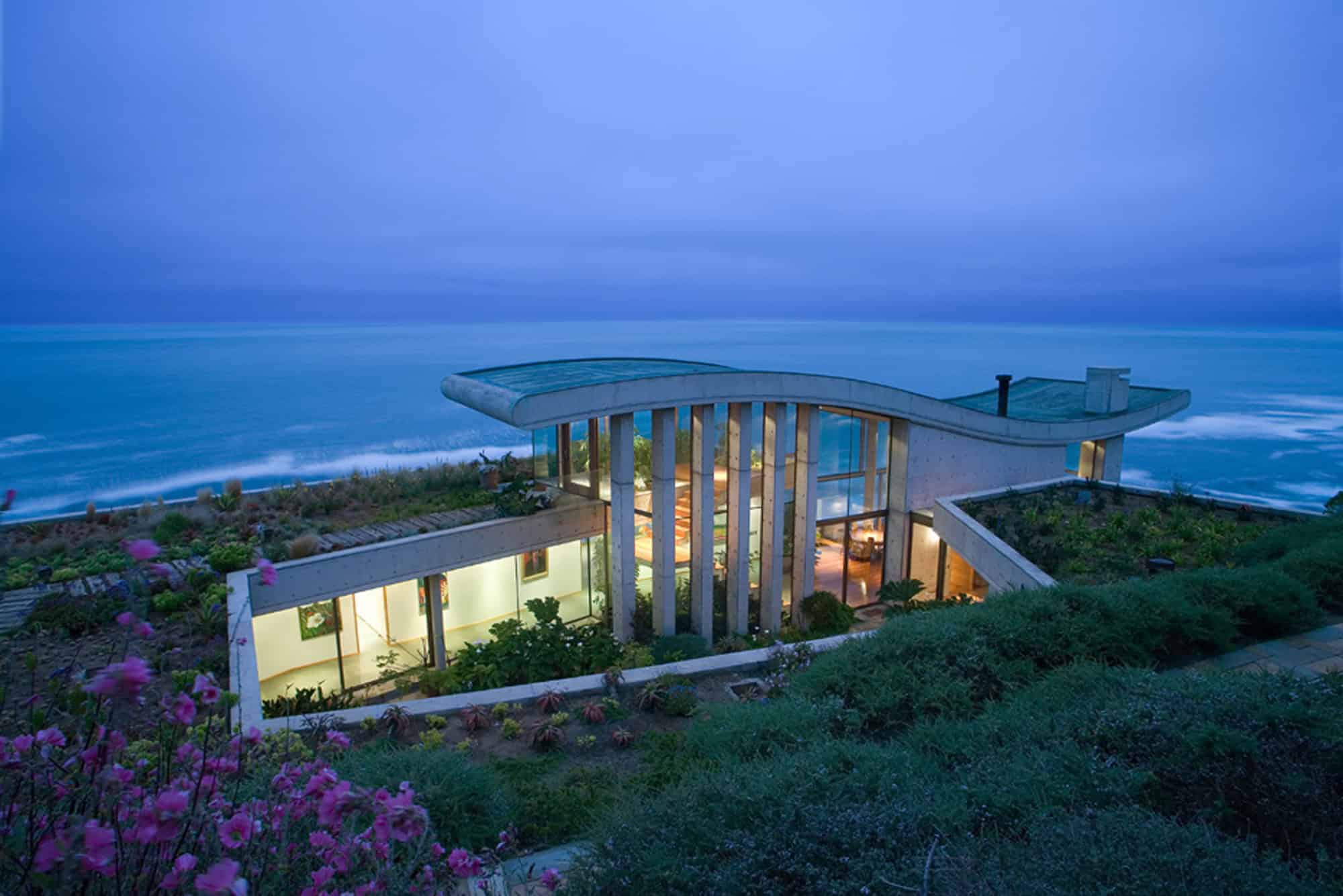 Modern beachfront house with curved roof and large glass windows, featuring innovative architectural design and lush landscaping by the ocean.