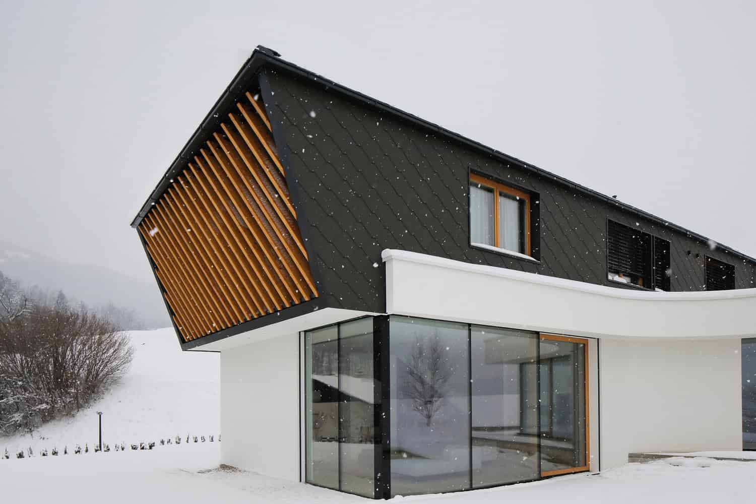 Contemporary modern house with unique architectural design, black exterior accents, large glass windows, and wooden features on a snowy landscape.