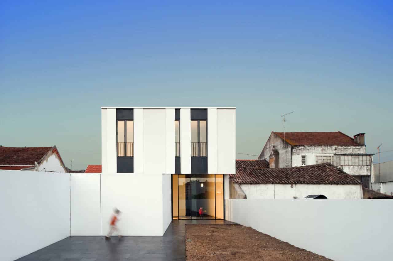 Modern minimalist house with clean white facade and large glass entryway, surrounded by a simple white boundary wall, blending contemporary architecture with traditional surrounding buildings.