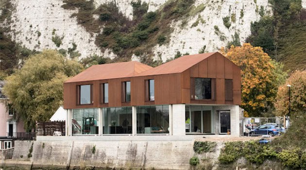 Modern waterfront house with glass facades and wooden accents, blending contemporary architecture with natural surroundings, set against a scenic hillside backdrop.