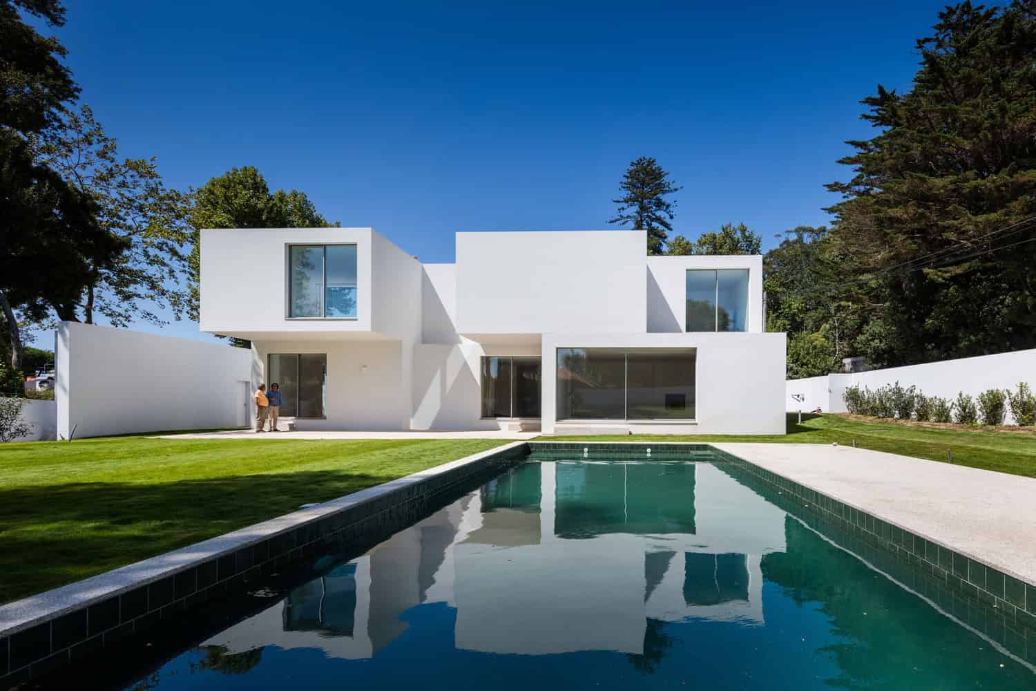 Modern white minimalist house with large glass windows and a sleek swimming pool in a spacious backyard, surrounded by lush greenery and tall trees, under a clear blue sky.