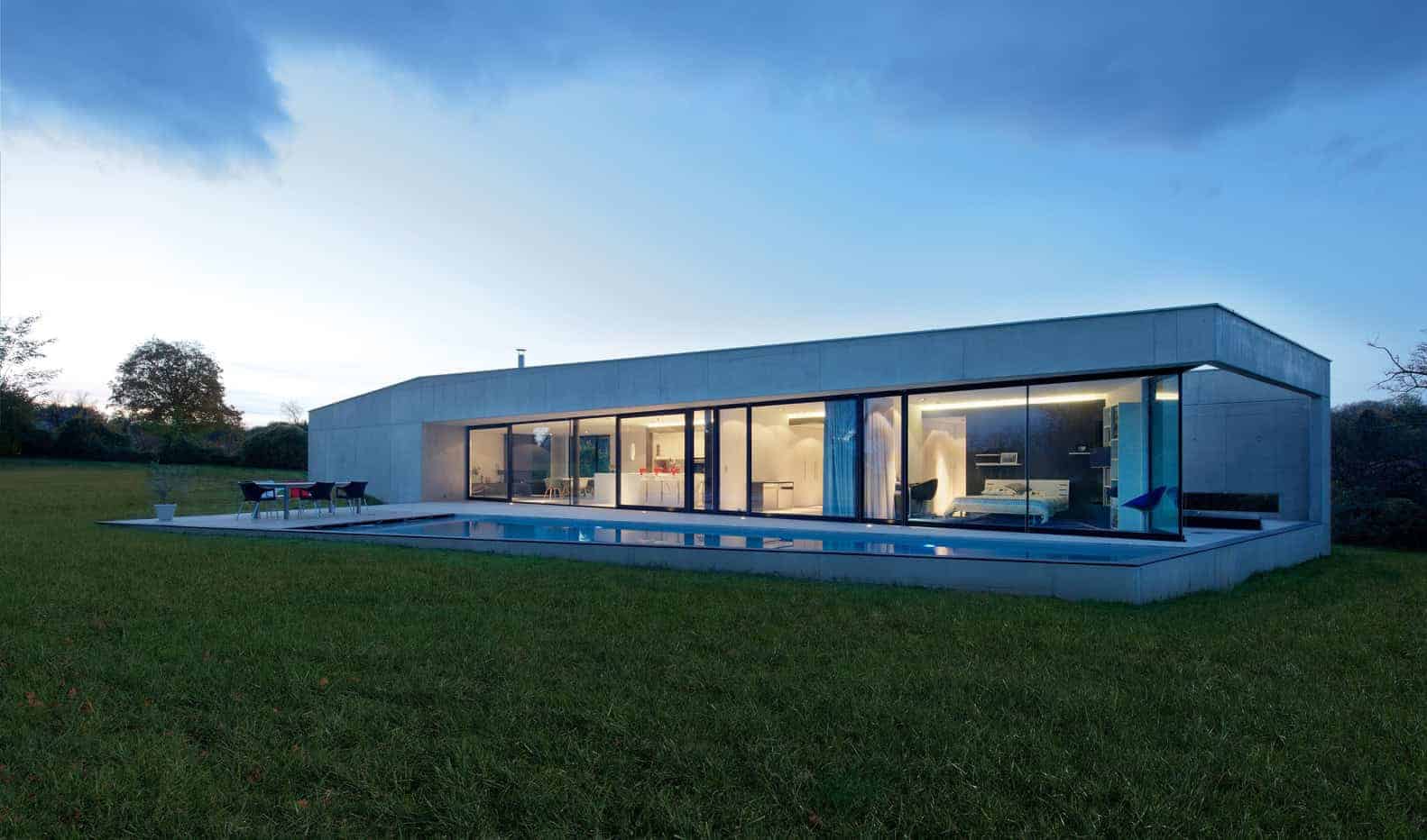 Sleek modern house with large glass walls and minimalist design, featuring an outdoor pool and lush green yard under a blue evening sky, exemplifying innovative architecture and contemporary home design.