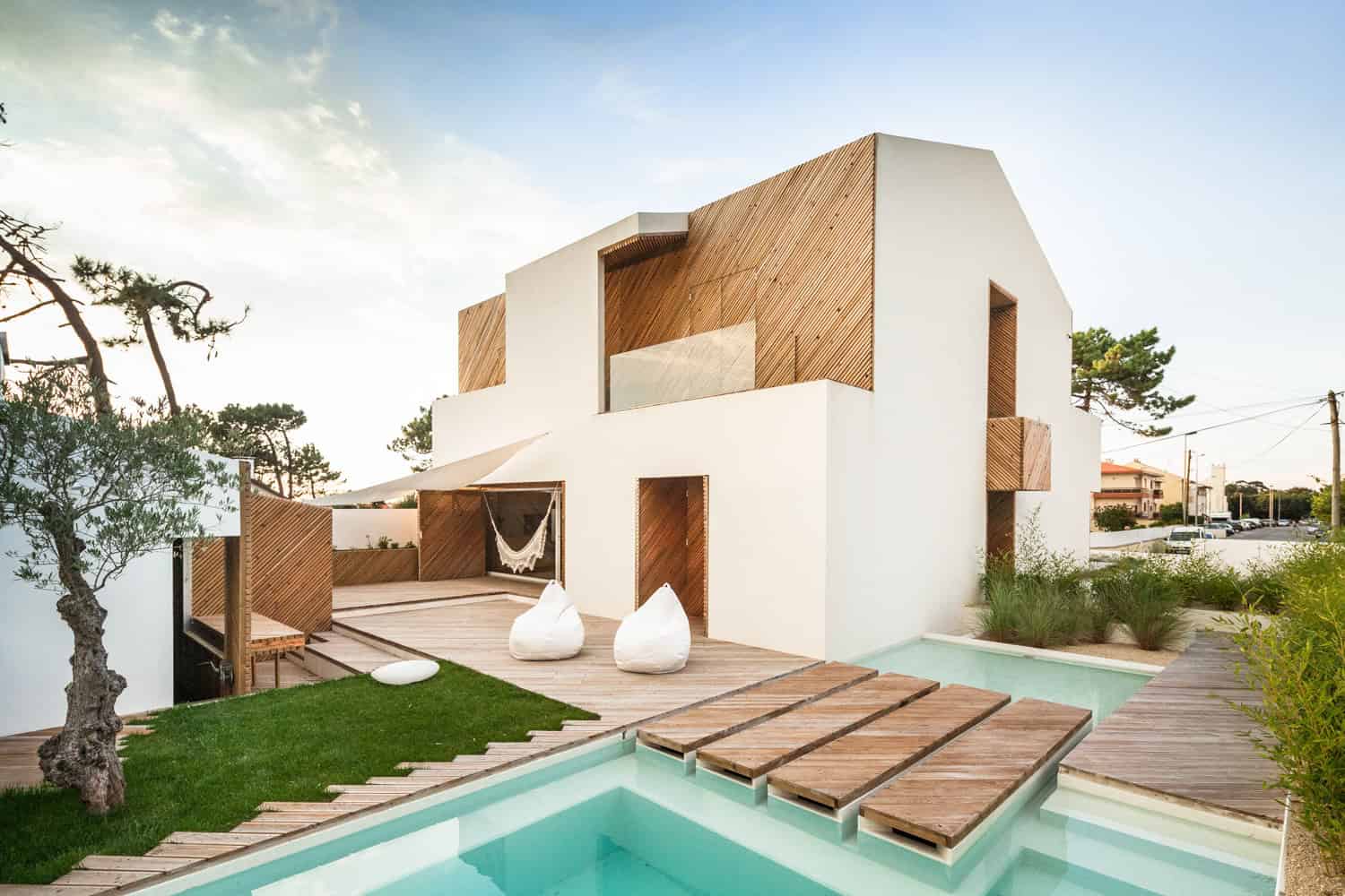 Modern minimalist house with white stucco walls and wooden accents, outdoor pool, and cozy seating area, surrounded by greenery and located in a sunny suburban neighborhood.