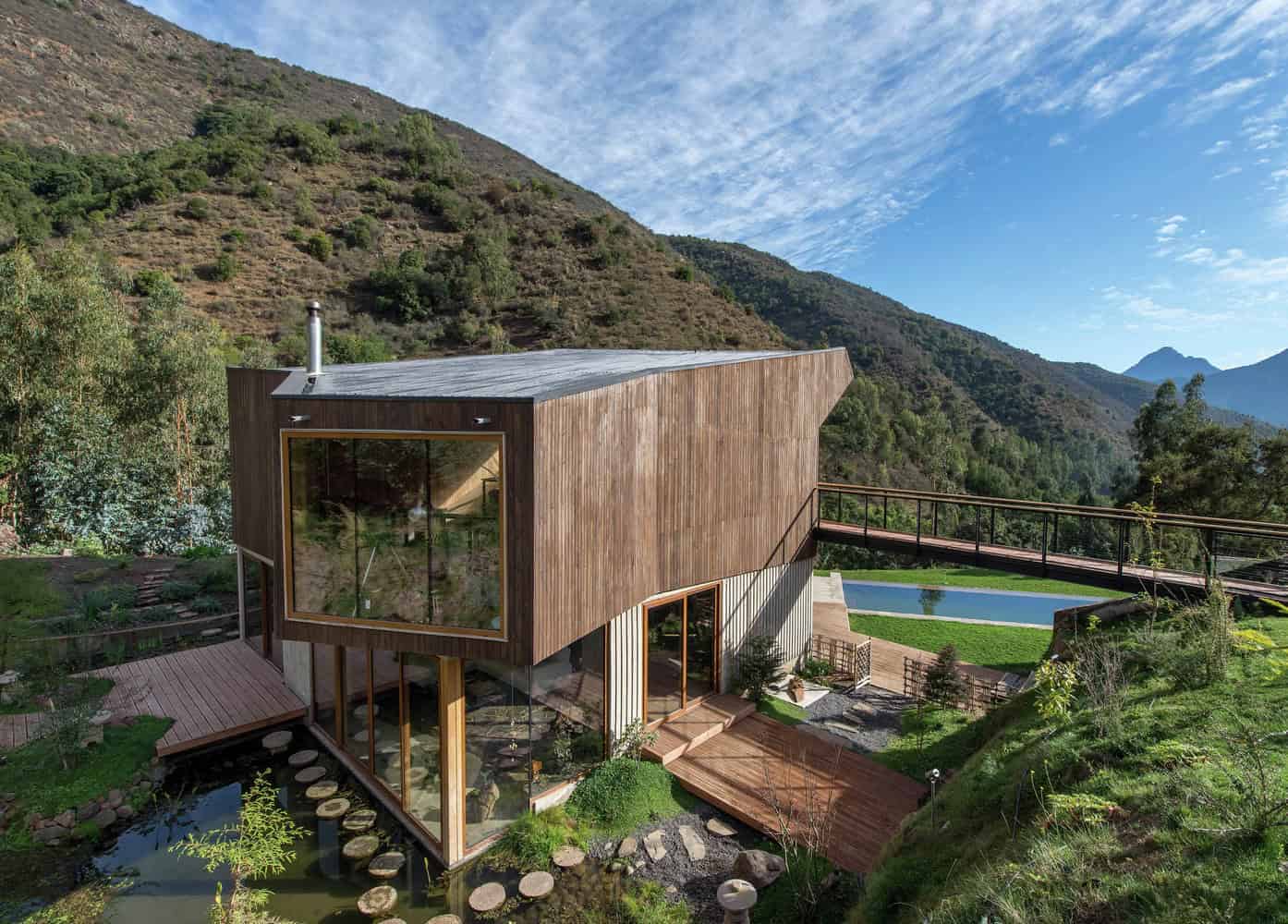 Contemporary mountain home with wooden exterior, large glass windows, and integrated natural landscape, set against lush green hills and blue sky. Elegant design blending modern architecture with nature.