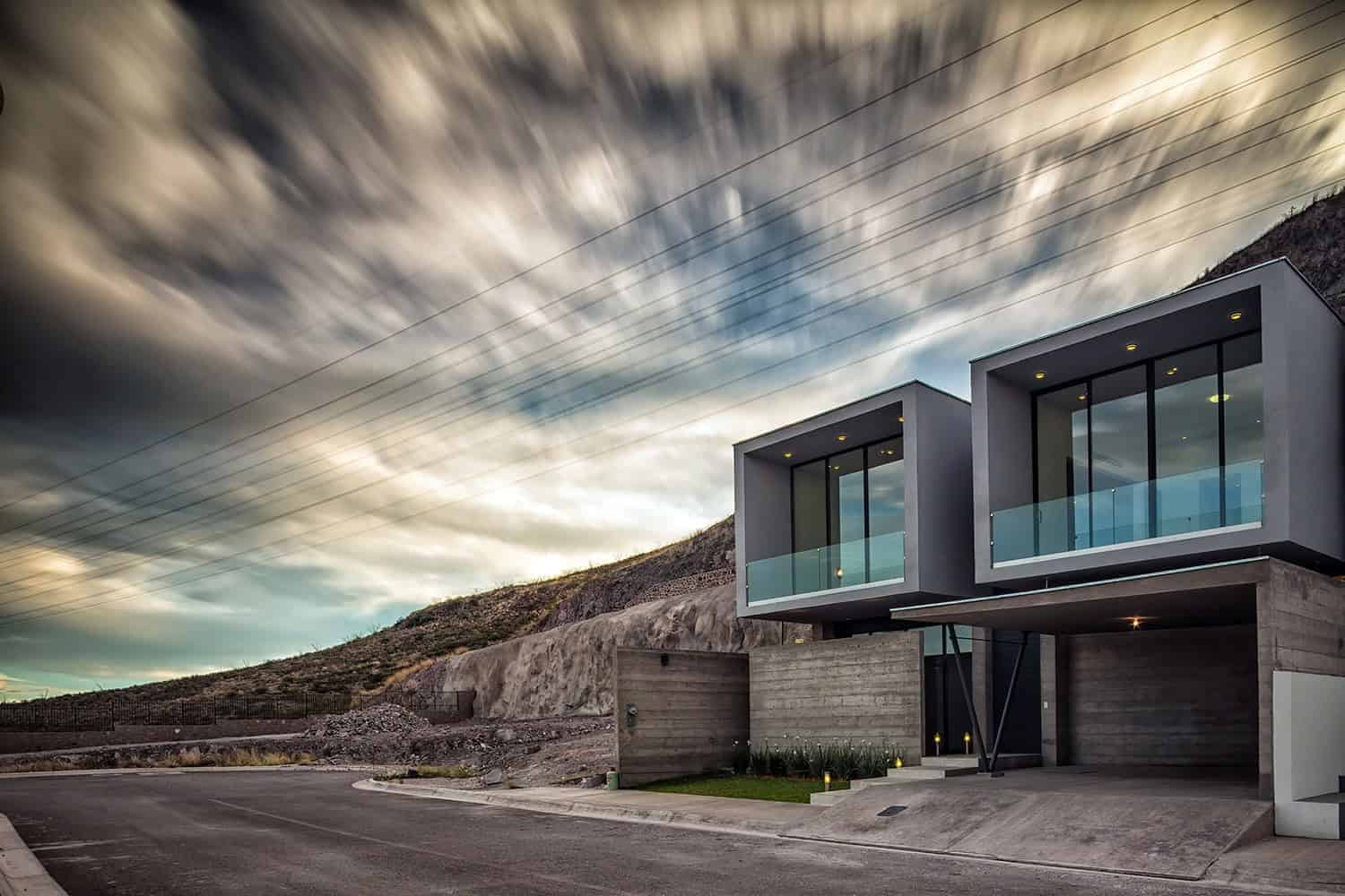Contemporary modern house with minimalist design and large glass windows on a hillside under dramatic cloudy sky.