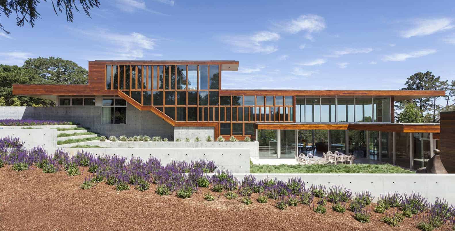 Modern architectural house with large glass windows and wooden accents, surrounded by landscaped garden and lavender plants. Contemporary home design with natural materials and spacious outdoor patio.
