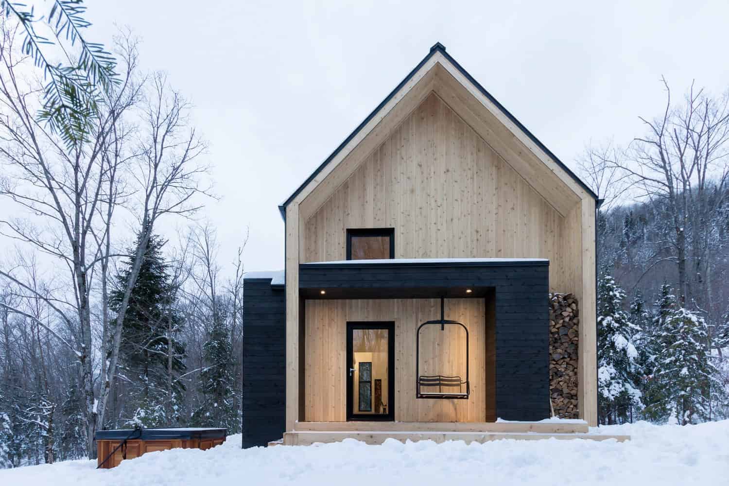Modern minimalist wooden house with black accents in snowy landscape, featuring a cozy outdoor swing and stacked firewood, showcasing contemporary architecture and winter design elements.