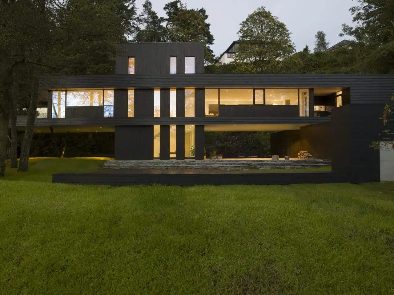 Modern black minimalist house with expansive glass windows and open floor plan, set amid lush greenery and trees, showcasing contemporary architecture and innovative design.