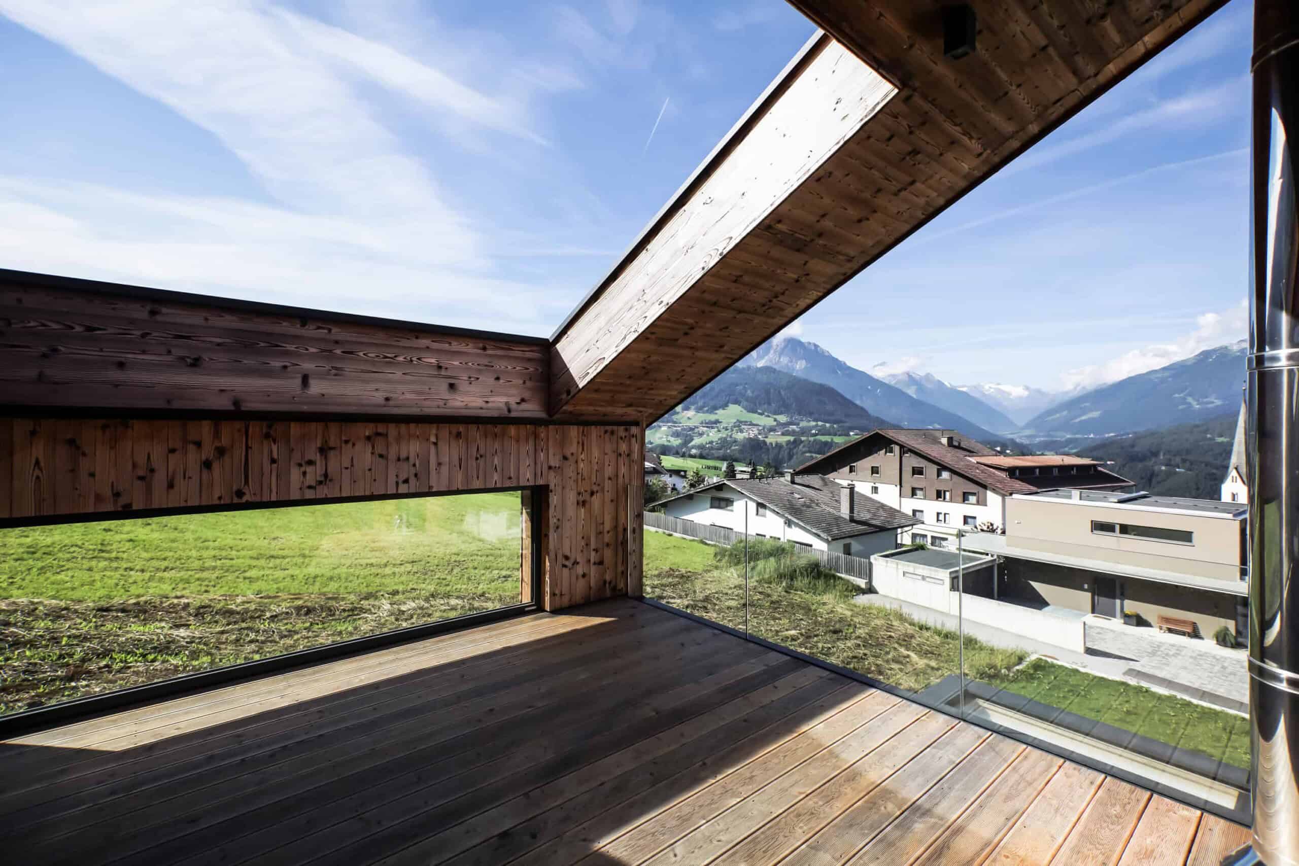 Modern mountain view balcony with glass railing and wooden flooring, showcasing scenic alpine landscape, mountains, and contemporary architecture in a picturesque rural setting.