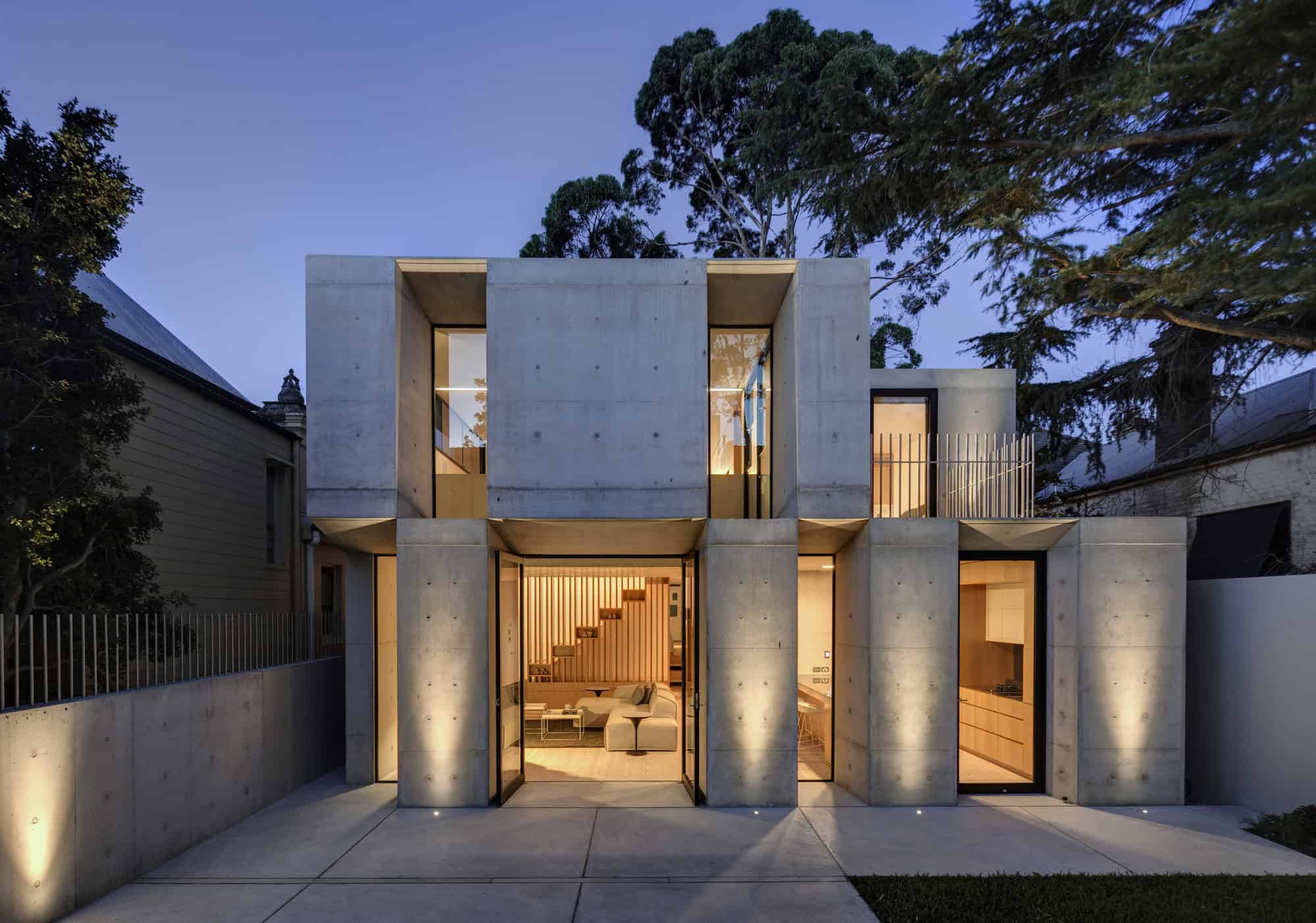 Contemporary concrete house exterior with large glass windows, modern architectural design, illuminated interior, surrounded by trees at dusk.