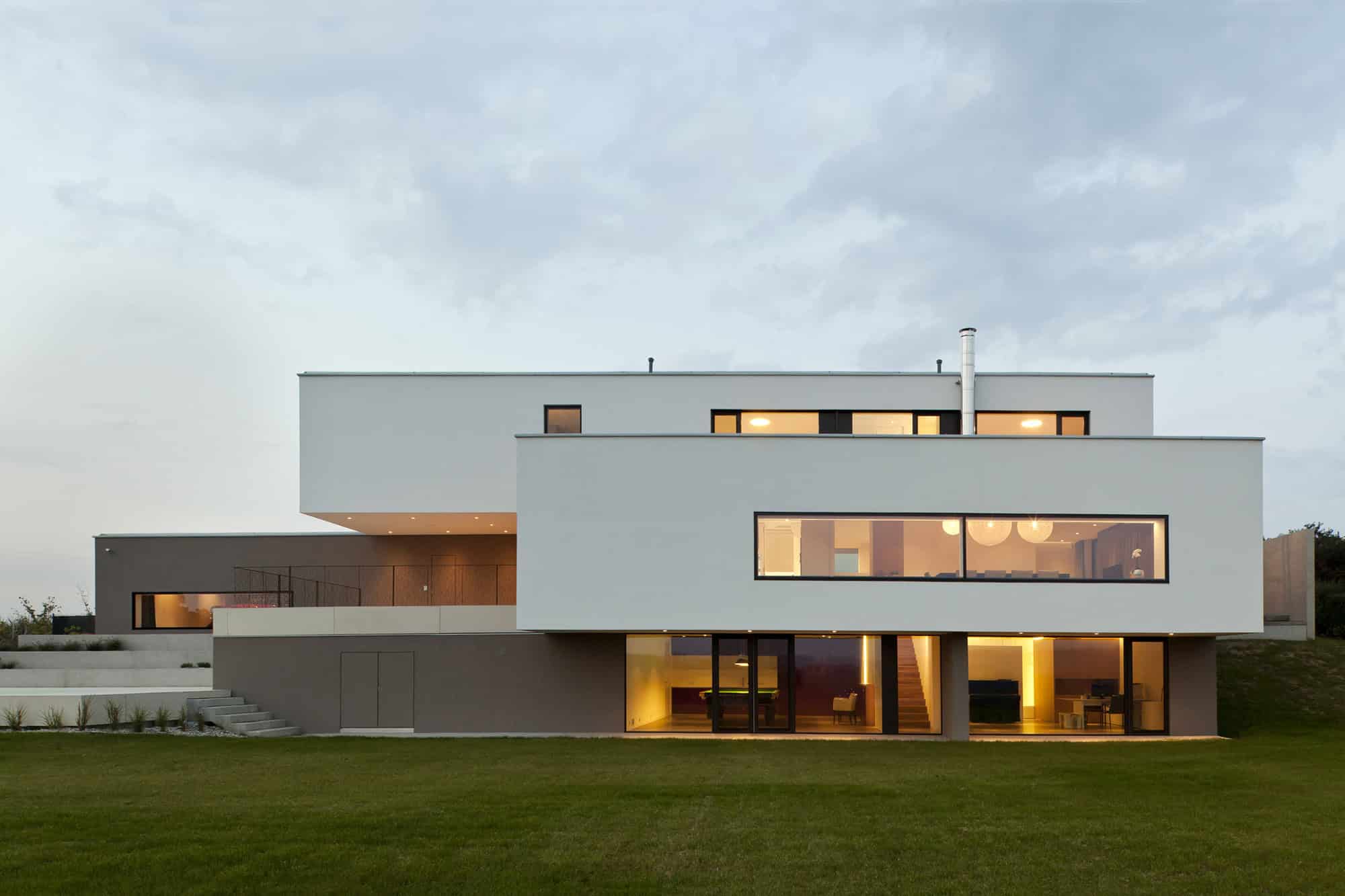 Contemporary modern house with minimalist architecture, large glass windows, and sleek geometric design, set on a landscaped lawn during dusk.
