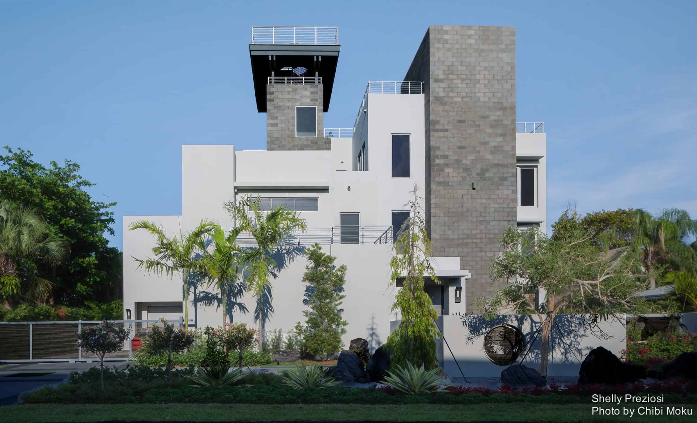 Modern minimalist house with clean lines, white facade, and contrasting gray brick accents, surrounded by lush landscaping and tropical plants.
