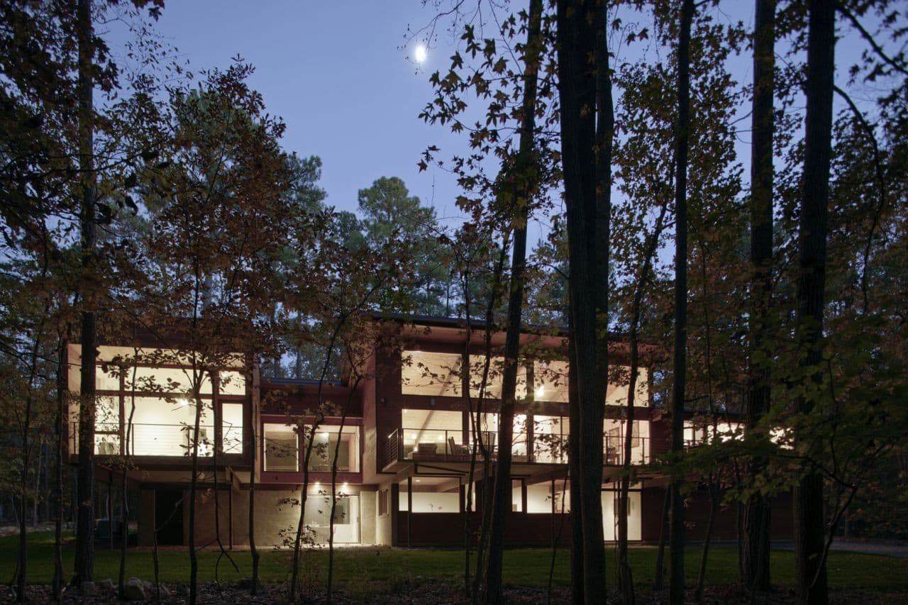 Modern contemporary house with large glass windows and illuminated interiors, surrounded by a forest during dusk, showcasing innovative architecture and unique design elements.
