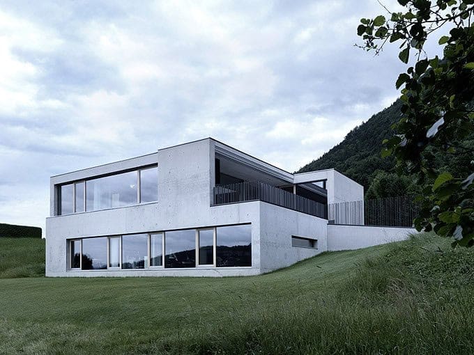 Modern minimalist concrete house with large glass windows overlooking green landscape and mountain backdrop, contemporary architecture design, luxury residential building in natural setting.