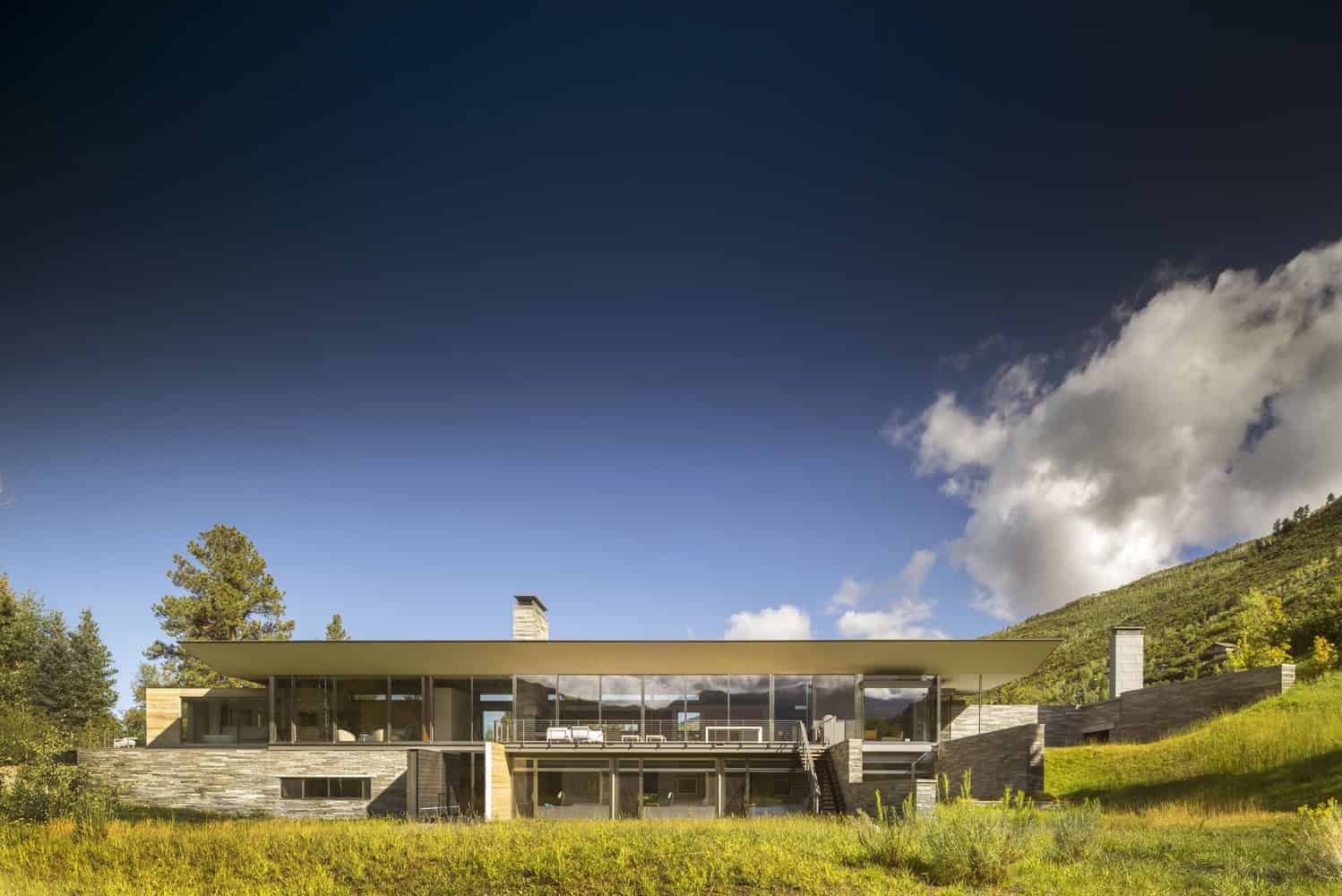 Contemporary modern house with expansive glass walls and flat roof set in a scenic hillside landscape.