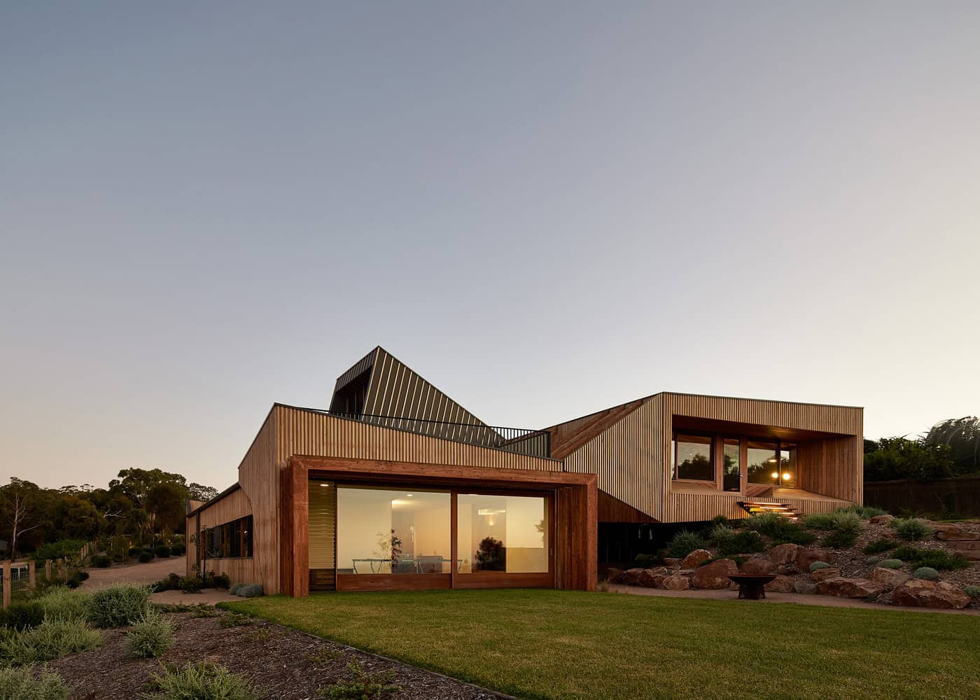 Contemporary wooden house with modern design and large glass windows on a landscaped lawn during sunset.