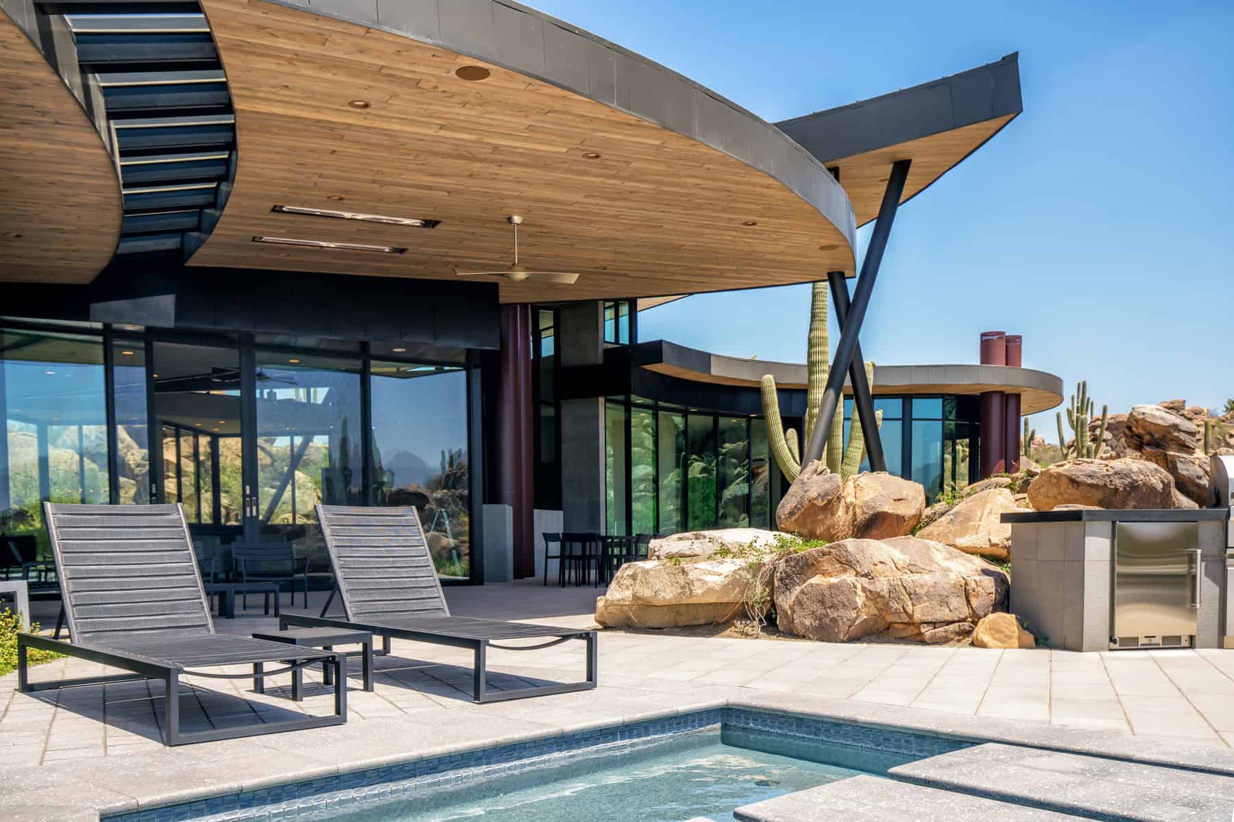 Modern desert-style house with an expansive outdoor patio, pool, and contemporary architectural design featuring wood elements, large glass windows, and desert landscaping with cacti.
