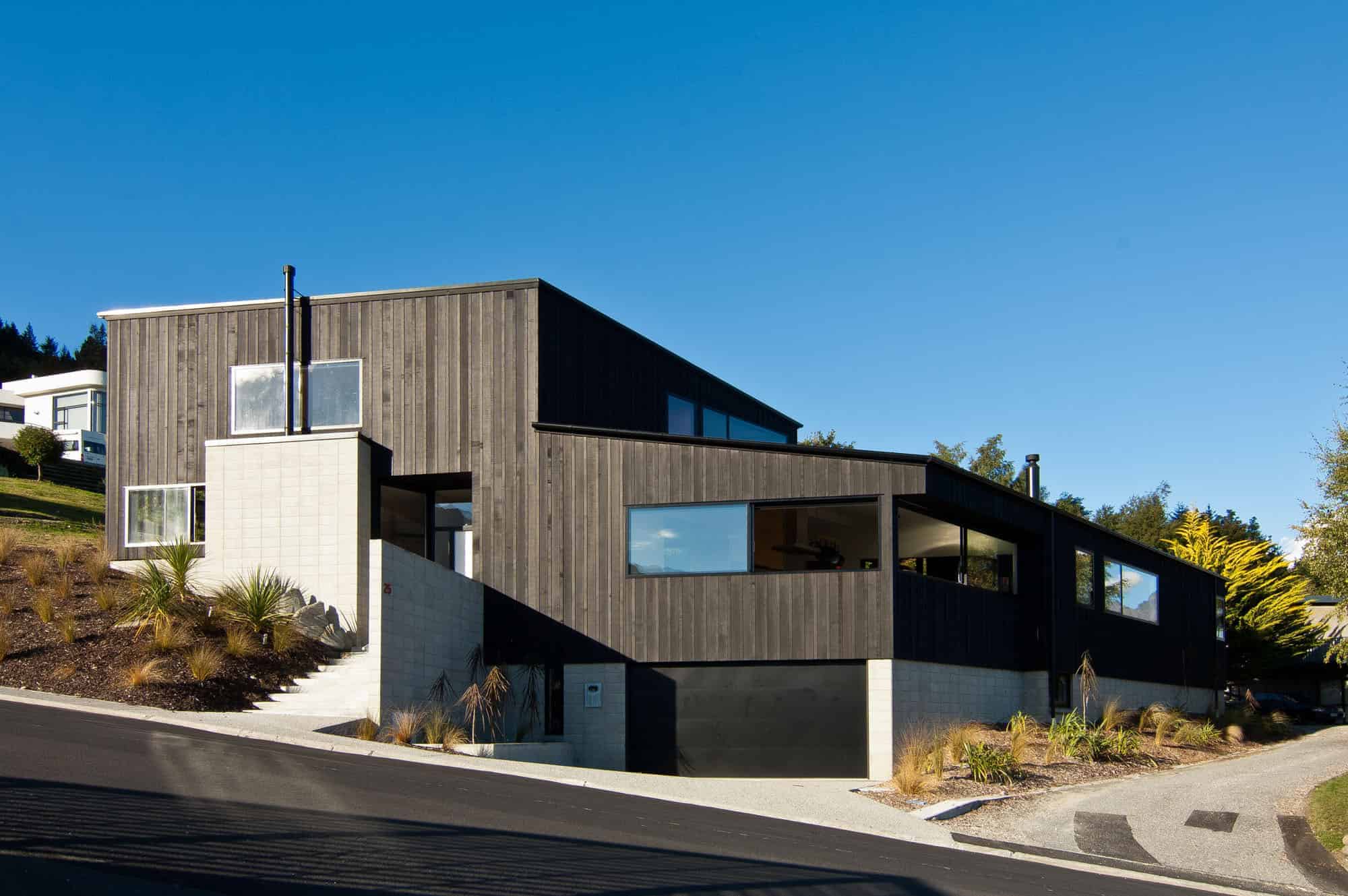 Contemporary modern house with dark wood exterior, large windows, and minimalist design on a sloped landscaped lot under a clear blue sky.