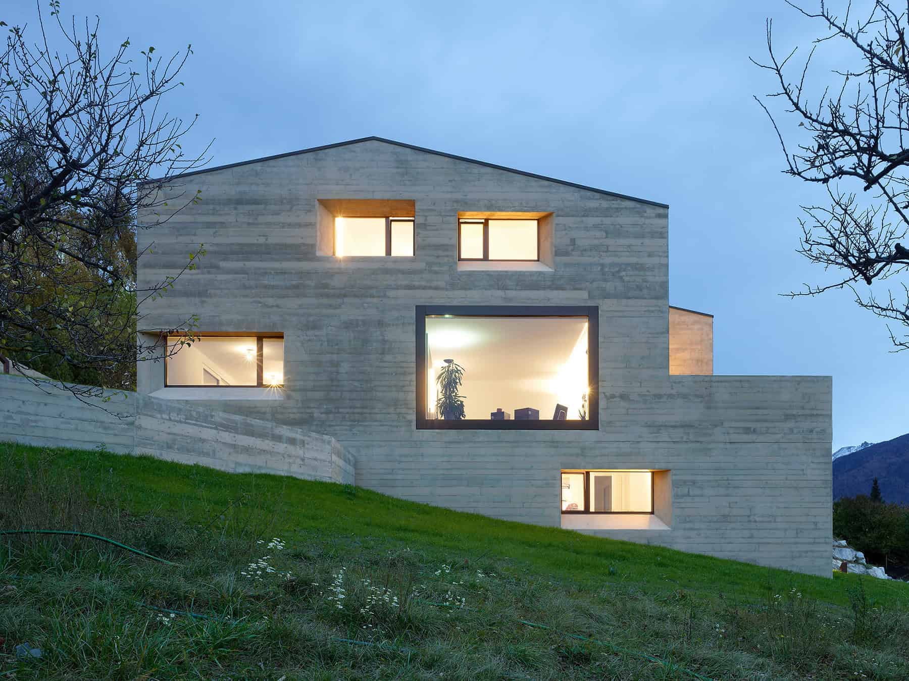 Contemporary minimalist concrete house with large windows, set on a grassy hillside in a scenic mountain landscape, showcasing modern architecture design.