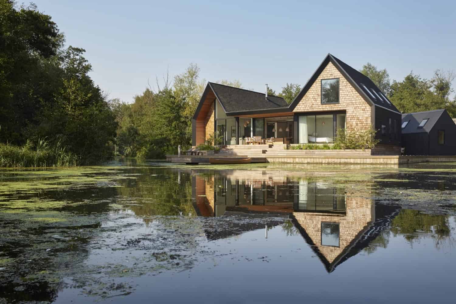 Contemporary modern lakeside house with large glass windows and wooden accents, reflecting in calm water surrounded by lush greenery and trees, showcasing innovative architecture and natural harmony.