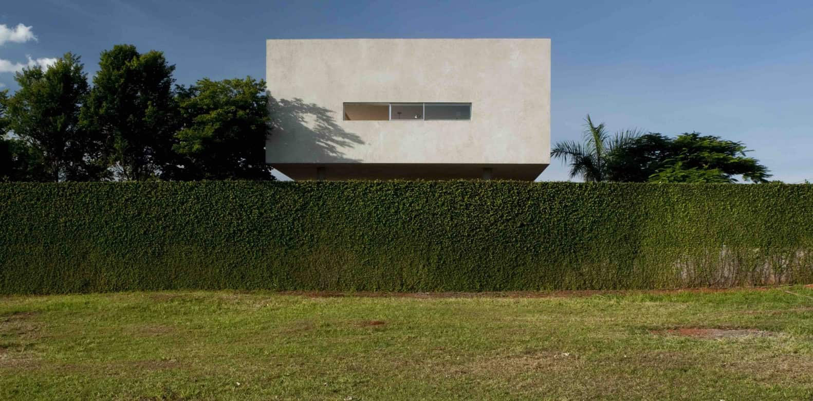 Modern minimalist concrete house with clean geometric lines and large horizontal window, surrounded by lush greenery and a well-manicured hedge, showcasing contemporary architecture design.