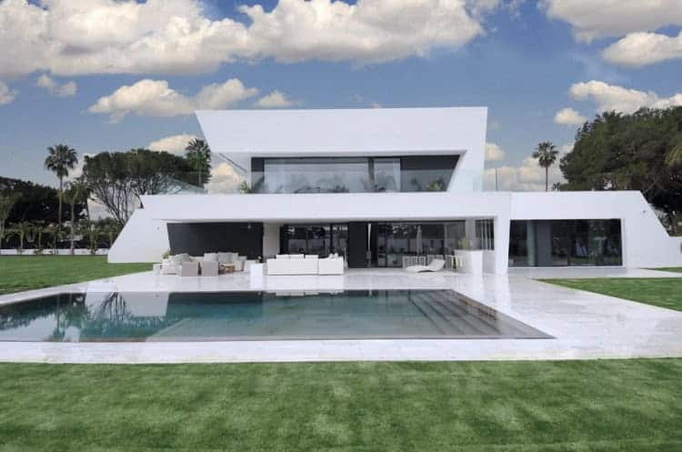 Modern luxury villa with contemporary architectural design and sleek white exterior. Features spacious outdoor patio, swimming pool, and lush green lawn. Perfect example of upscale residential architecture.
