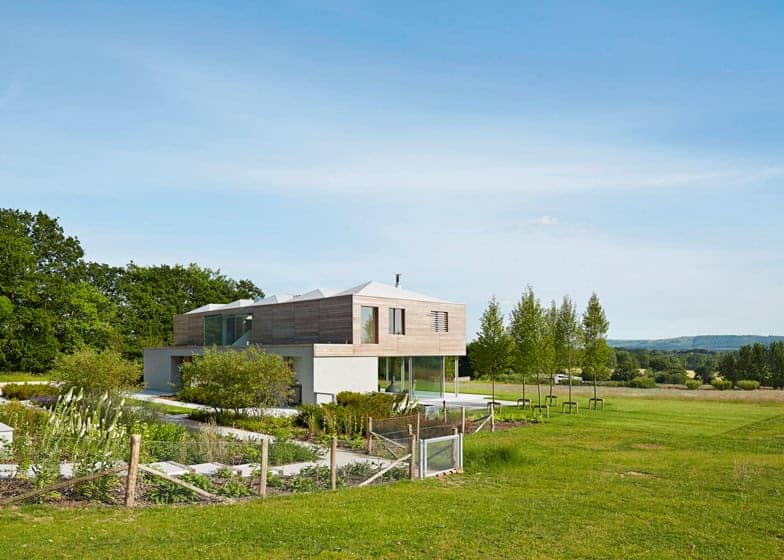 Modern minimalist house with sleek wooden exterior and large glass windows, situated in a lush green landscape with open fields and clear blue sky.