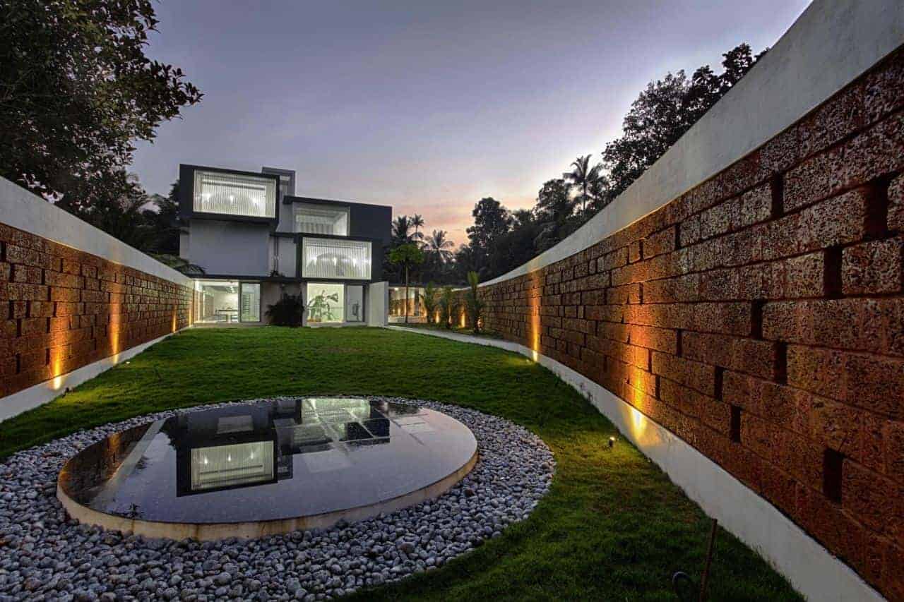 Modern contemporary house with unique cubic architecture illuminated at dusk, featuring sleek glass elements, lush greenery, and a reflective pond, showcasing innovative design and sophisticated landscape architecture.