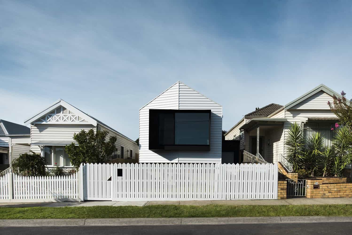 Modern minimalist house with bold black window frame and white siding, surrounded by a white picket fence and lush greenery, showcasing contemporary architecture and home design.