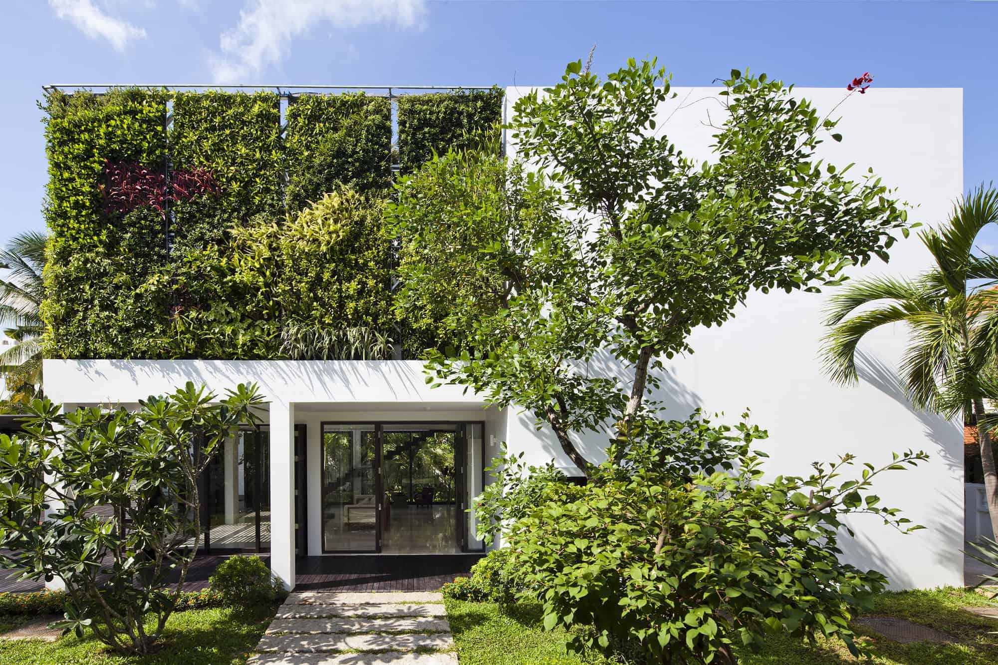 Lush green modern residential building with vertical garden and minimalist white facade, surrounded by tropical plants and trees, featuring sleek glass doors and contemporary landscaping.
