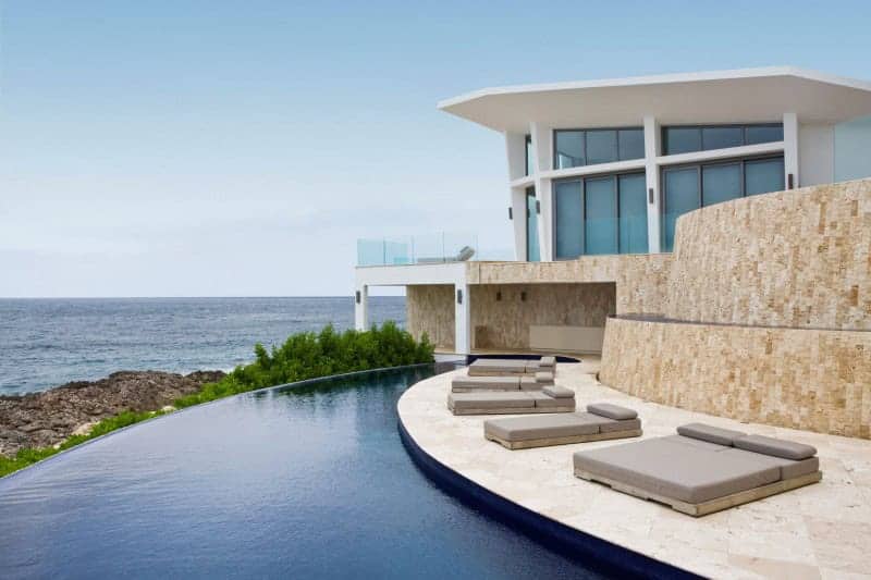 Modern luxury beachfront house with infinity pool and ocean view, featuring contemporary architecture, large glass windows, and outdoor lounging area.