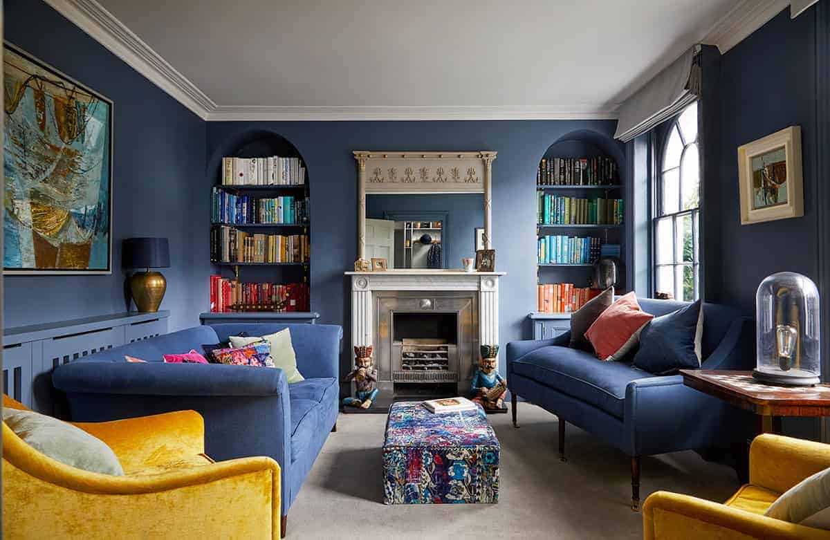 Luxurious living room with navy blue walls, elegant fireplace, colorful velvet sofas, and built-in bookshelves filled with books, creating a cozy and stylish ambiance.