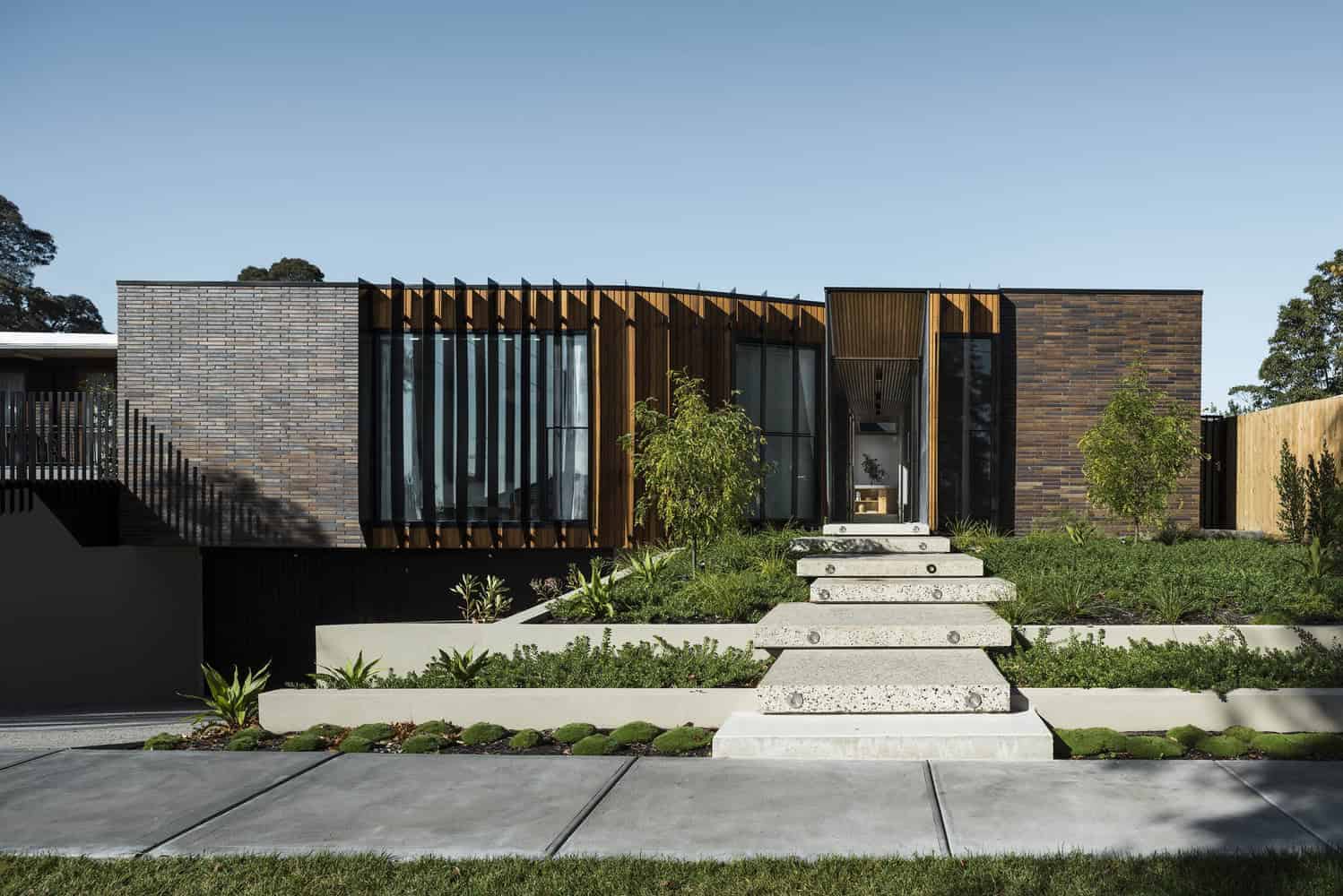 Contemporary modern house with unique architectural design, featuring brick walls, wooden accents, and large glass windows, set amidst lush landscaped greenery and a clean concrete pathway.